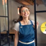Portrait of positive business woman standing at cafeteria door entrance. Cheerful young waitress in blue apron near glass door with open signboard and looking at camera. Excited small business owner.